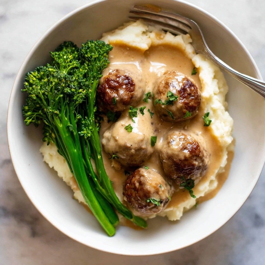 A white bowl holds a dish with three main layers, starting from the bottom with creamy mashed potatoes in an off-white color and soft texture. On top of the mashed potatoes are six brown meatballs, some covered fully in light brown sauce and others partially showing a slightly crispy, browned surface. The sauce is creamy and smooth, blanketing the meatballs and pooling in the bowl. To the right side of the bowl, bright green broccolini with small, textured florets and long stems sits, adding a fresh contrast to the dish. A silver fork is placed on the left side of the bowl, resting on the mashed potatoes and meatballs. The bowl is set on a white marbled surface. photo taken with an iphone --ar 1:1 --v 7
